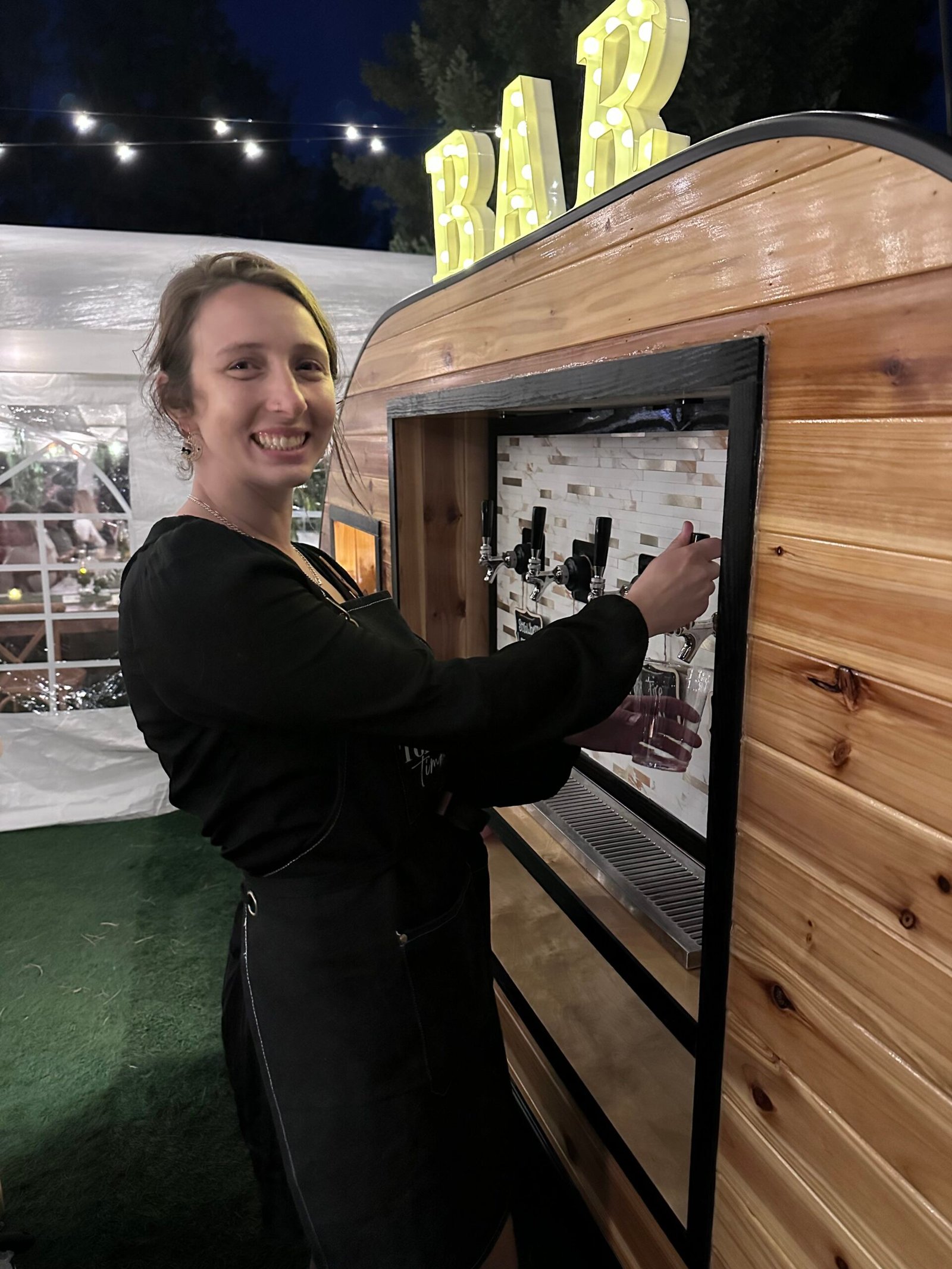 Kaley, owner of Tonic and Timber, smiling while pouring a drink from the tap trailer at a nighttime event